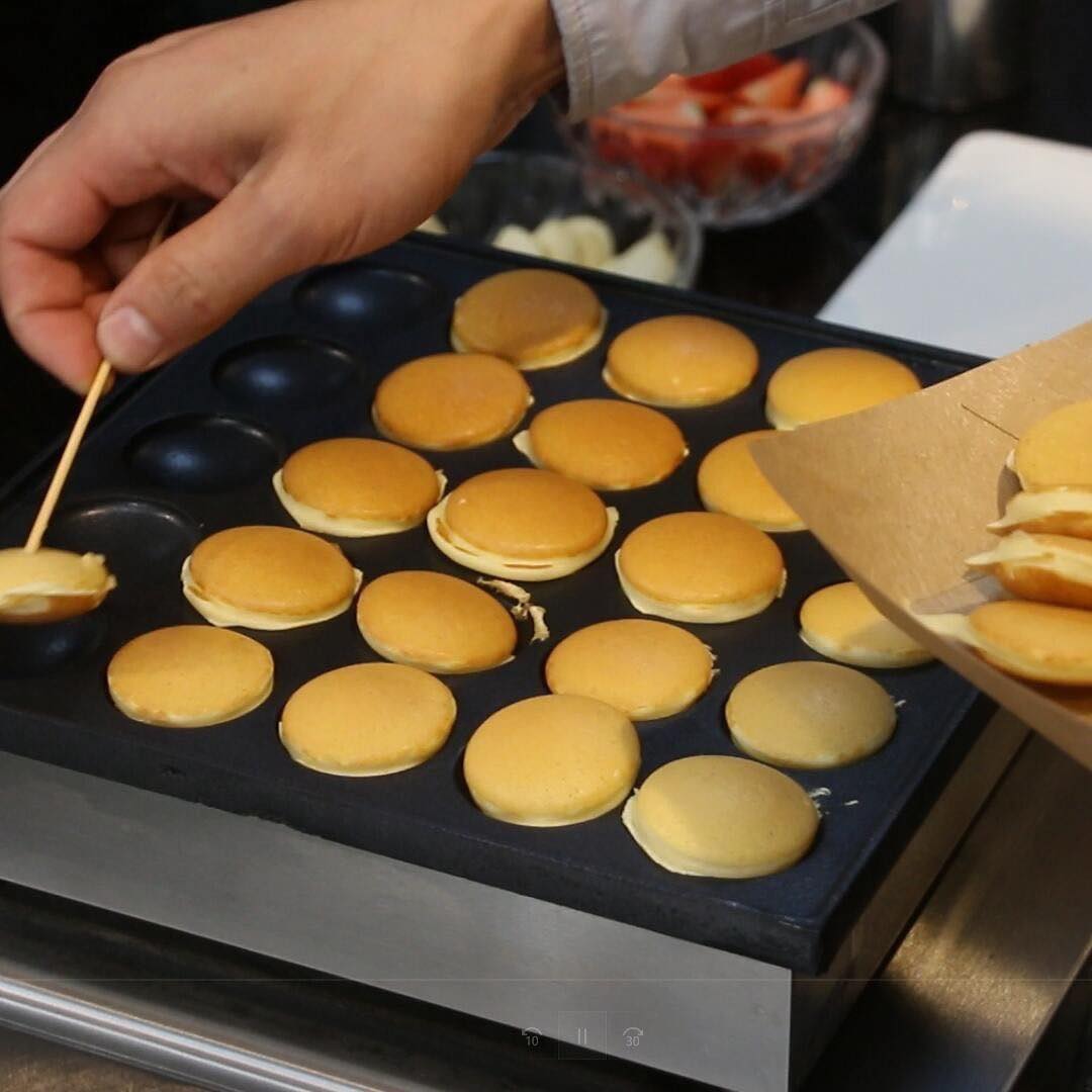 Mano cocinando mini panqueques dorados en la plancha, con fresas y banano de fondo